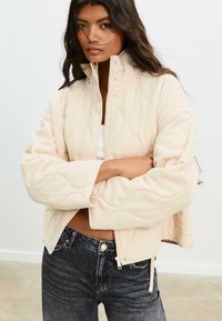 Lys beige quiltet jakke med høj krave, lynlåslukning og sidelommer. Sat sammen med mørk denim jeans. Glat tekstur.