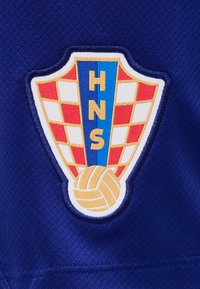 Κροατικό ποδοσφαιρικό έμβλημα με κόκκινο και λευκό καρό, μπλε κάθετη λωρίδα με τα γράμματα HNS και κίτρινη μπάλα ποδοσφαίρου στο κάτω μέρος σε μπλε ύφασμα.