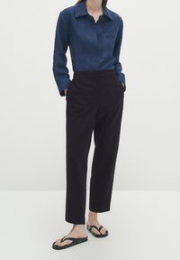 Denim shirt met een puntige kraag, lange mouwen en een borstzak. Gecombineerd met donkere tapered broeken en zwarte slippers.