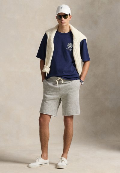 Μπλε μπλουζάκι, γκρι σορτς, λευκό πλεκτό γιλέκο που είναι περασμένο πάνω από τους ώμους, λευκά αθλητικά παπούτσια και λευκό καπέλο με λογότυπο. Μαύρα γυαλιά ηλίου.