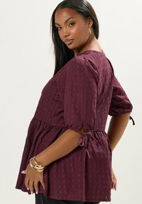 Top peplum bordeaux in tessuto texturizzato con maniche a sbuffo corte e dettagli con lacci ai lati. Presenta una vestibilità morbida e un motivo sottile.