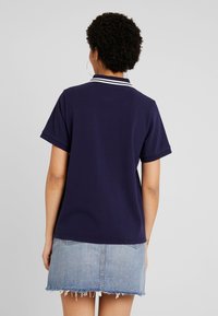 Femme aux cheveux bouclés portant des créoles, un polo bleu marine avec des rayures blanches sur le col, et une mini-jupe en jean effilochée, vue de dos.