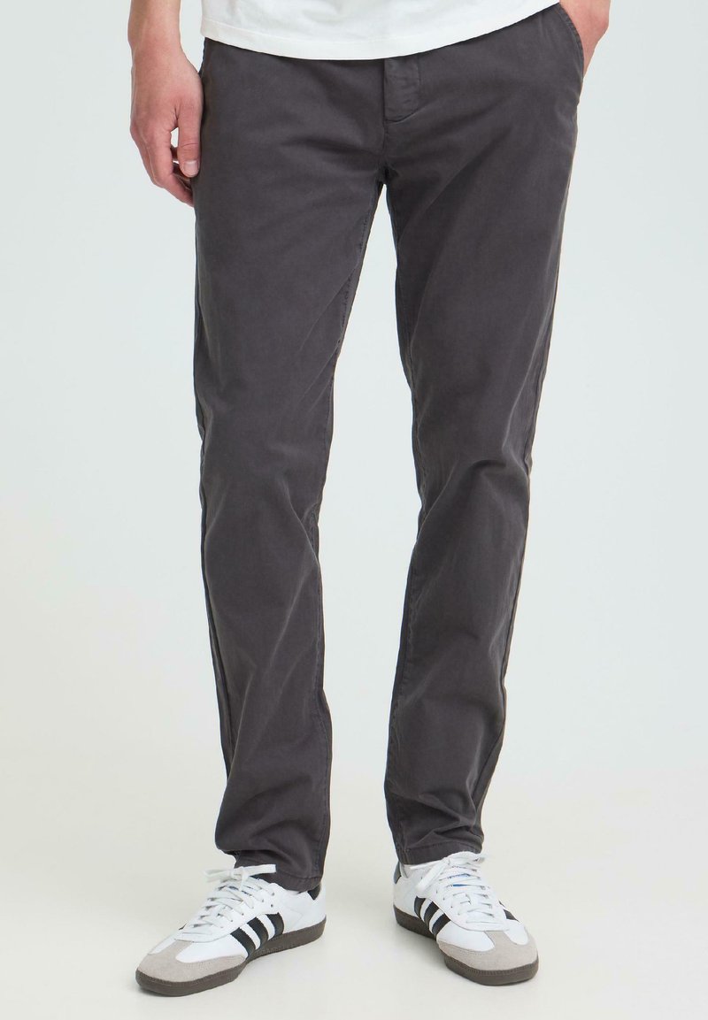 Homme portant un pantalon slim gris foncé et des baskets blanches avec des rayures noires, debout les mains dans les poches devant un fond uni.