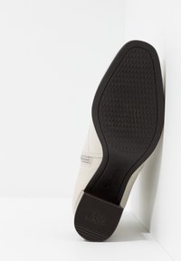Chaussure beige clair avec une tige lisse et une semelle extérieure en caoutchouc noir texturée. Présente un design subtil avec des lignes horizontales près du dessus.