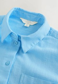 Chemise en lin bleu clair avec une texture douce, col pointu et patte de boutonnage. Comprend une poche poitrine et un motif tissé subtil.