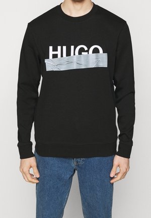 Mand iført sort sweatshirt med hvid tekst "HUGO", delvist dækket af grå duct tape-design, kombineret med blå jeans.