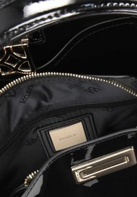Intérieur d'un sac à main en cuir verni noir montrant une fermeture éclair dorée, une doublure en tissu de marque et une étiquette métallique avec le logo "kazar" à l'intérieur de la poche.