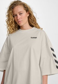 Overmaat beige t-shirt gemaakt van zachte katoen. Voorzien van een zwart afgedrukt "hummel sportswear" logo en zwarte chevron strepen op de mouwen.