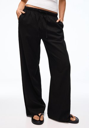 Personne portant un pantalon noir ample avec taille élastique et cordon, un top court blanc et des sandales noires, mains dans les poches.