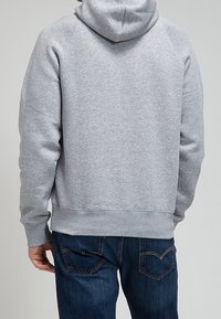 Personne portant un sweat à capuche gris clair et un jean bleu foncé, vue de dos sur un fond uni gris clair.