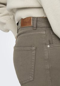 Suéter de punto beige combinado con jeans topo, con una etiqueta de cuero marrón que dice "el denim azul" y detalles de botones metálicos en los bolsillos.