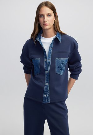 Felpa blu navy con accenti in denim, caratterizzata da una chiusura frontale con bottoni, due tasche sul petto e una vestibilità comoda. Texture morbida e dettagli a contrasto.