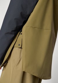 Giacca verde oliva con una sezione nera sulla spalla, caratterizzata da una texture liscia, una chiusura a scatto e un design a vestibilità ampia.