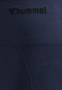 Close-up af mørkeblåt tekstureret stof med ribbede sektioner og et sort "hummel" logo og biesymbol øverst i midten.