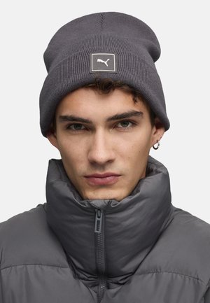 Gray knitted beanie with a branded patch; soft texture. The model wears a high-collar gray puffer jacket, showing a neutral color palette.