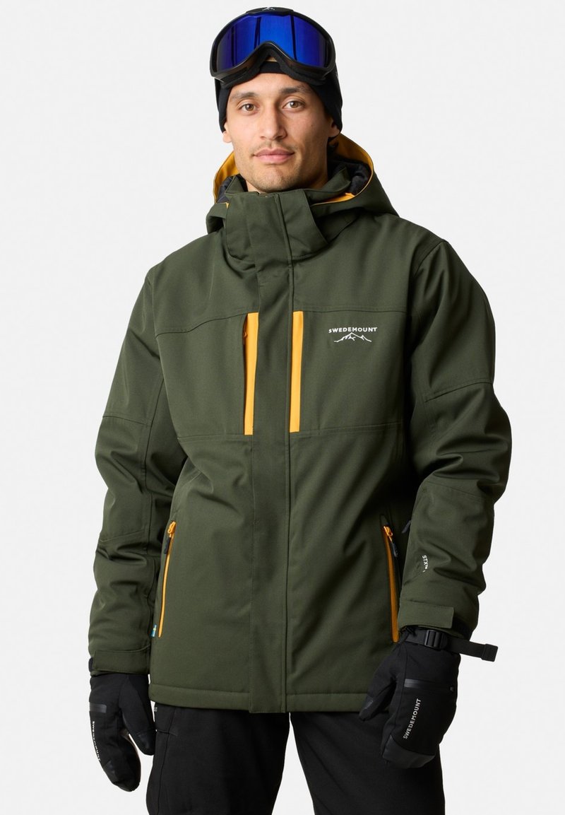 Giacca da sci verde con accenti arancioni, tasche con zip, cappuccio e polsini regolabili. Indossata con guanti neri e occhiali da sci.