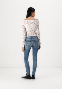 Fehér virágmintás, hosszú ujjú felső, vállnélküli kivitelben, kék skinny farmernadrággal és fekete magas szárú tornacipővel párosítva, hátulról nézve.