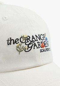 Gorra de algodón blanca con texto bordado "el JARDÍN NARANJA" y diseños florales en amarillo, rojo y azul. Textura suave con visera curvada.