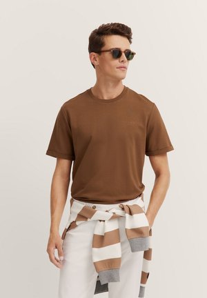 Brown cotton t-shirt with a crew neck and short sleeves. Worn with white pants and a striped sweater tied at the waist. Round sunglasses.
