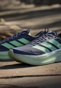 Μπλε και πράσινα παπούτσια τρεξίματος Adidas με ανοιχτοπράσινες σόλες με την επιγραφή Dreamstrike Glow σε πλακόστρωτη υπαίθρια επιφάνεια με φυσικό φως.