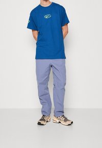 Modrá bavlněná trička s žlutým logem Nike, doplněná o světlé fialové kalhoty a béžové tenisky s černými akcenty a síťovaným designem.