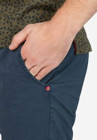 Marineblaue Shorts mit glatter Textur, ausgestattet mit einer Seitentasche und einem roten Logoakzent. Der Stoff ist leicht und im lässigen Design.