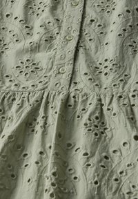 Blusa di cotone verde chiaro con intricate ricami floreali, chiusure a bottoni e una texture arricciata vicino all'orlo.