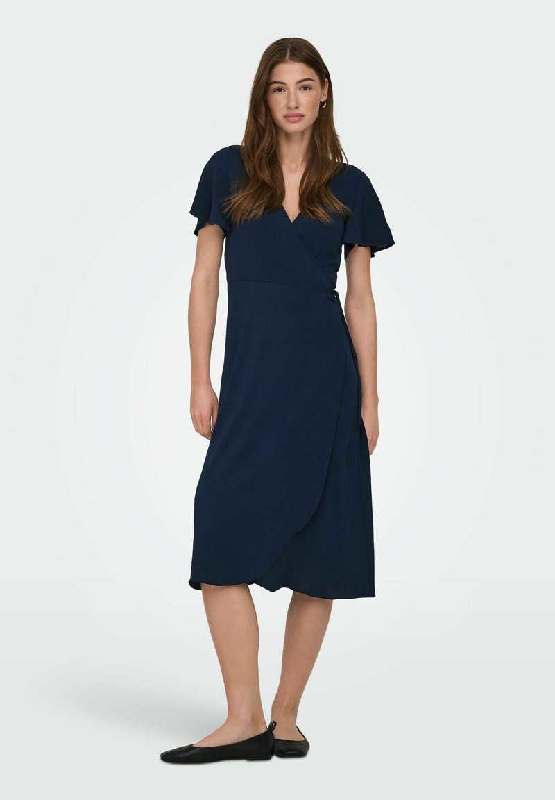 Femme aux longs cheveux bruns portant une robe portefeuille bleu marine et des ballerines noires, debout devant un fond clair uni.