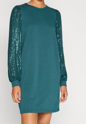 Długa sukienka z długim rękawem w kolorze zielonkawoniebieskim, z cekinowymi, teksturowanymi bufkami oraz dopasowanymi mankietami, noszona przez osobę o opalonej skórze i naturalnych paznokciach.