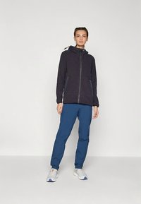 Ciemna bluza polarowa z zamkiem z przodu, regulowanym kapturem i bocznymi kieszeniami, połączona z niebieskimi spodniami i białymi trampkami. Gładka tekstura.