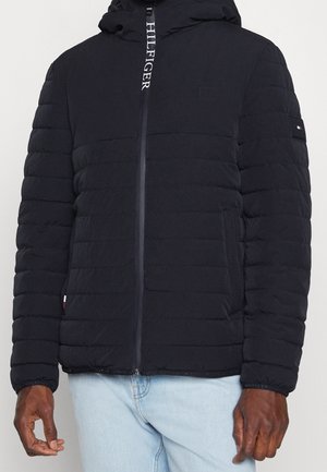 Czarna pikowana kurtka z kapturem, suwak z przodu z napisem "HILFIGER", noszona na jasnoniebieskich dżinsach.