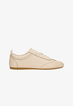 Sneaker in pelle beige con profilo basso, punta rotonda, suola piatta, cuciture sottili e allacciatura frontale. Design minimalista senza loghi visibili.