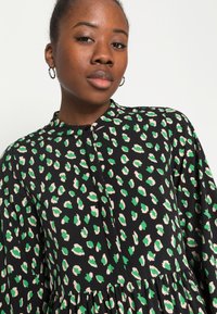 Vestido de tecido preto com padrão de leopardo em verde e rosa pálido, colarinho alto, fecho de botão e detalhe de saia franzida.