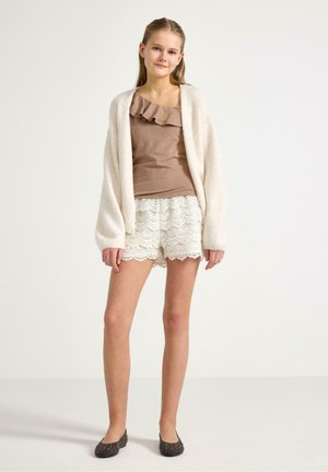 Nuori tyttö seisoo, pukeutuneena ruskeaan röyhelöiseen yläosaan, valkoisiin pitsishortseihin, kermaan väriseen neuletakkiin ja tummiin niitattuihin balleriinoihin, taustana yksivärinen pinta.