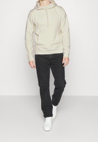 Beżowy hoodie z przednią kieszenią i kapturem na sznurki, w połączeniu z czarnymi dżinsami i białymi sneakersami. Prosty design z minimalnymi akcentami.
