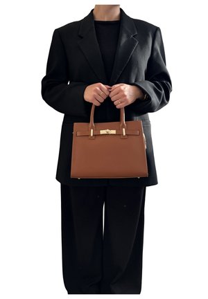 Persona in abito nero che tiene una borsa di pelle marrone con hardware dorato, centrata su uno sfondo bianco.