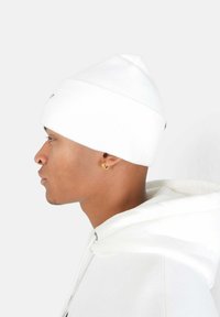 Vit stickad mössa med en avsmalnande form, utrustad med en vikbar kant och diskret logotypdetalj. Matchad med en vit huva-sweatshirt.