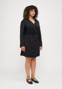 Vestito in wrap nero con maniche lunghe, scollo a V, dettaglio con fiocco in vita e tessuto morbido; abbinato a scarpe nere flat.