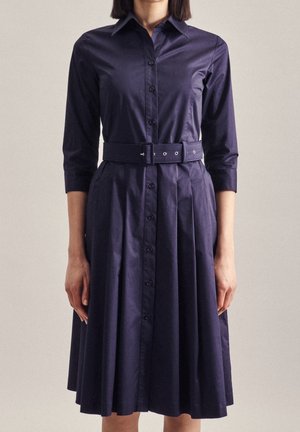 Kobieta ubrana w granatową sukienkę-płaszcz na guziki z rękawami do łokci i szerokim, dopasowanym paskiem w talii, stojąca na jednolitym tle.