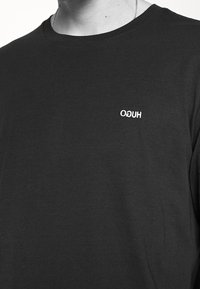 Schwarzes Langarm-T-Shirt aus Baumwolle, mit einem weißen gestickten "HUGO"-Logo auf der oberen linken Brust. Glatte Textur, Rundhalsausschnitt-Design.