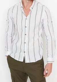 Valkoinen pitkähihainen paita, jossa ohuet ruskeat pystysuorat raidat. Edessä napillinen, hihansuut käännetyt. Valmistettu kevyestä kankaasta.