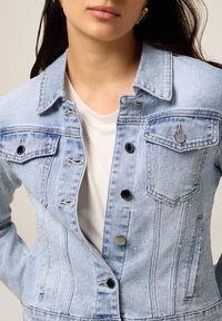 Veste en denim bleu clair avec un devant à boutons, deux poches poitrine et un motif texturé subtil avec des gouttes, agrémentée de boutons noirs.