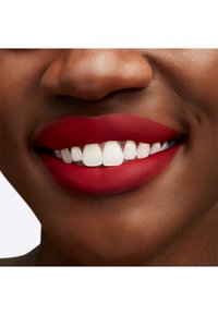 Couleur à lèvres rouge mate sur des lèvres pulpeuses, contrastant avec une peau lisse et claire. Dents blanches éclatantes visibles, soulignant un sourire sain.