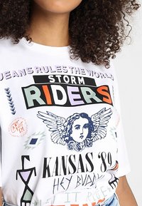 Bílý tričko s barevným textem "STORM RIDERS," grafikou anděla s křídly, "KANSAS '89," a různými abstraktními symboly a kresbami.