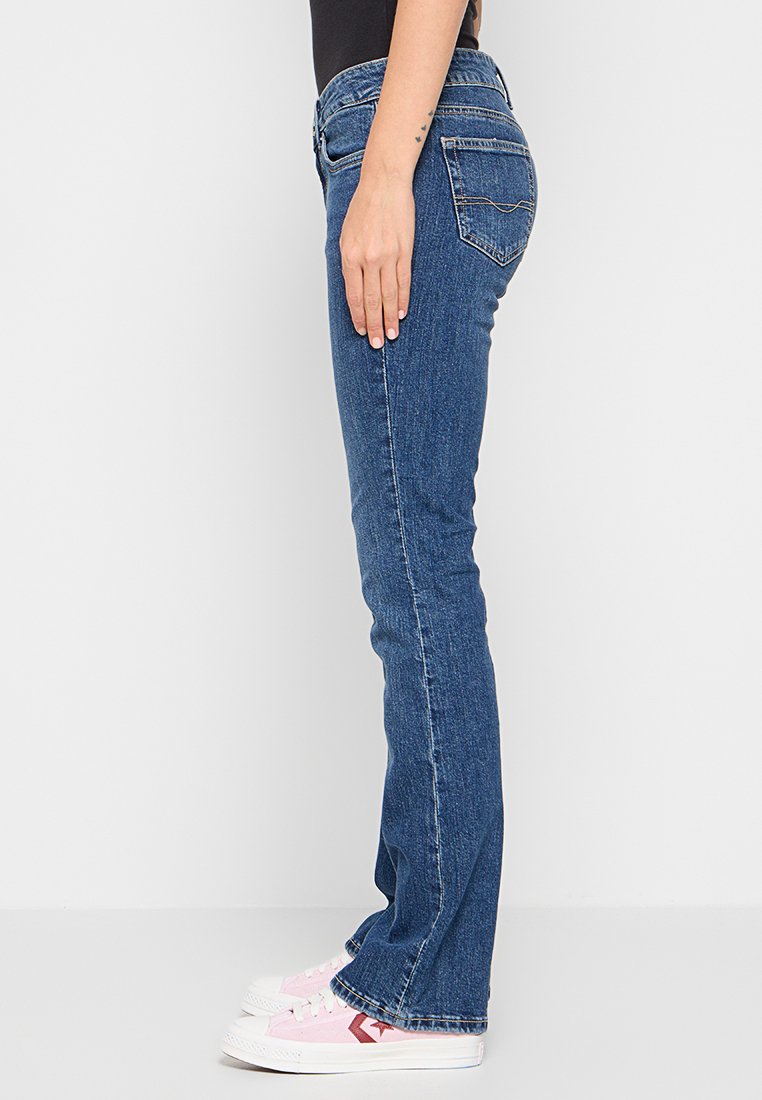 Ausgestellte Denim-Jeans in Dunkelblau mit einem mittelhohen Bund, fünf Taschen und dezenten Nähten. Kombiniert mit pinken Sneakers.