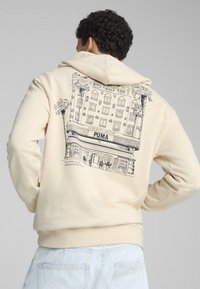 Felpa beige con grafica sulla schiena che presenta il contorno di un edificio, palme e il testo "PUMA", mettendo in risalto una texture liscia.