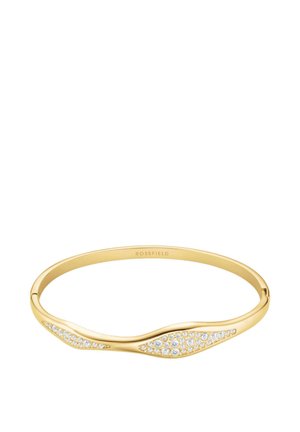 Pulsera tipo brazalete en tono dorado con diseño ondulado, presenta cristales transparentes incrustados en un lado, acabado pulido y marca discreta en el interior.