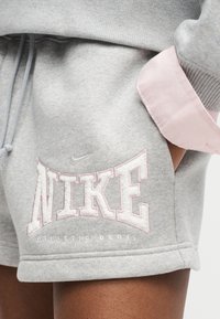 Κοντινό πλάνο γκρι σορτς Nike Athletic Dept. που φοριούνται με το χέρι στην τσέπη και στρωμένα πάνω από μανσέτα ανοιχτό ροζ πουκαμίσου.