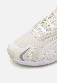 Lyst beige atletisk sko med en tekstureret mesh-overdel, hvide snørebånd og gummisål. Har grå accenter og perforerede detaljer.