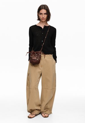 Kobieta w czarnym swetrze na guziki, luźnych beżowych spodniach typu cargo, brązowej szydełkowanej torbie na ramię i brązowych sandałach, stojąca na białym tle.